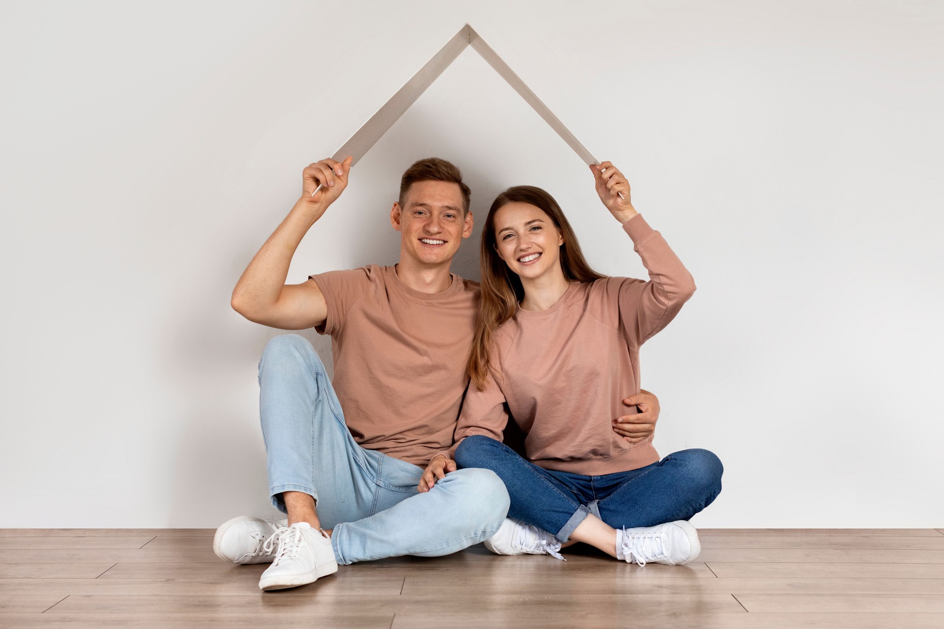 Real Estate. Happy Couple Sitting Under Symbolic Cardboard Roof Near White Wall